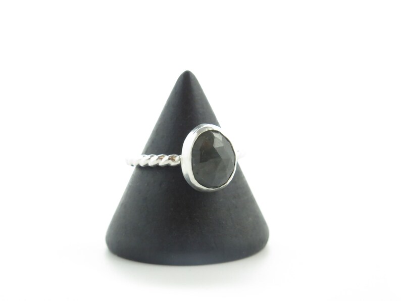 K&ouml;nnte beinhalten: Ein silberner Ring mit einem gedrehten Band und einem dunkelgrauen Edelstein. Der Ring wird auf einem schwarzen kegelf&ouml;rmigen St&auml;nder pr&auml;sentiert.