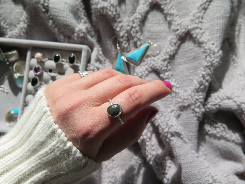 K&ouml;nnte beinhalten: Ein silberner Ring mit einem dunkelgrauen Edelstein, der in einer Zargenfassung gefasst ist. Der Ring befindet sich an einem Finger.