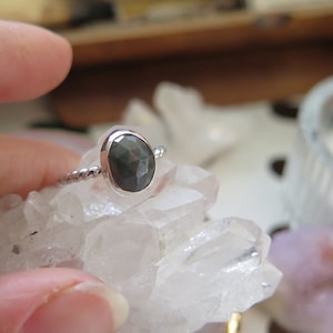K&ouml;nnte beinhalten: Ein silberner Ring mit einem dunkelgrauen ovalen Edelstein, der in einer Zargenfassung sitzt. Der Ring liegt auf einem Bett aus wei&szlig;en Kristallen.