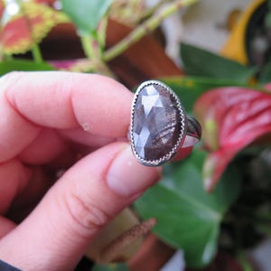 Può includere: Un anello in argento con una grande pietra preziosa marrone scuro sfaccettata a forma di pera, incastonata in una lunetta dentellata. La fascia dell'anello presenta un motivo testurizzato e inciso sulla superficie, offrendo un contrasto.