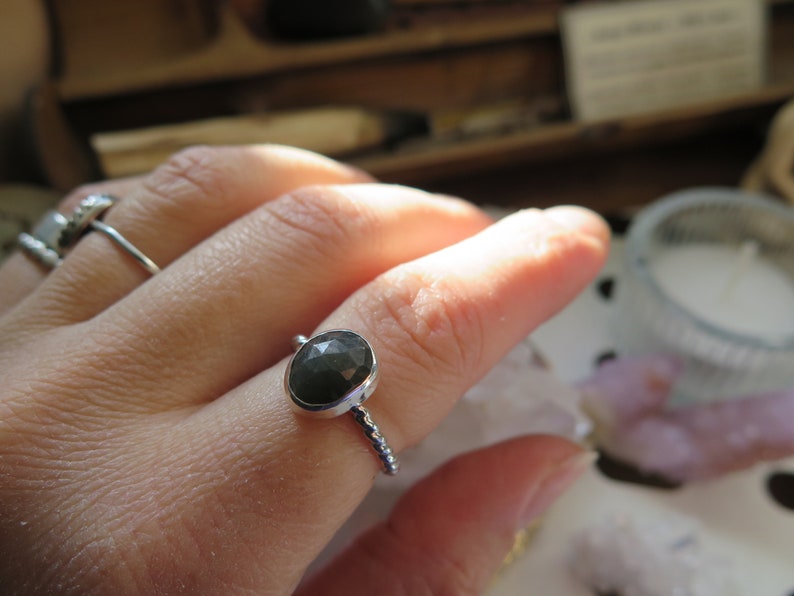K&ouml;nnte beinhalten: Ein silberner Ring mit einem dunkelgrauen Edelstein, der in einer Zargenfassung gefasst ist. Der Ring hat eine strukturierte Band.