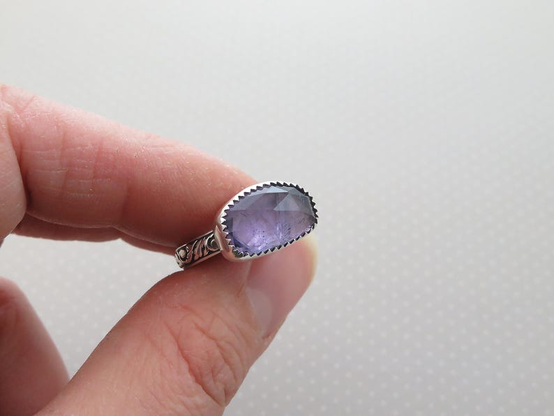 May include: A silver ring with a large, rectangular, faceted amethyst gemstone. The ring has a detailed, ornate band.