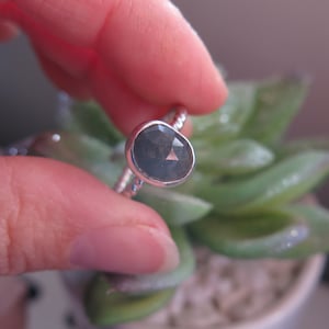 K&ouml;nnte beinhalten: Ein silberner Ring mit einem dunkelgrauen ovalen Edelstein. Der Ring ist auf einem strukturierten Band gesetzt.