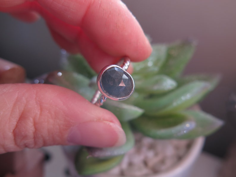 K&ouml;nnte beinhalten: Ein silberner Ring mit einem dunkelgrauen ovalen Edelstein. Der Ring ist auf einem strukturierten Band gesetzt.