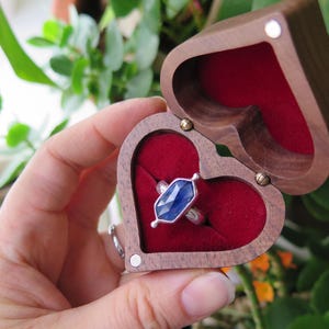 Op de afbeelding: Een ring met een blauwe edelsteen in een hartvormige houten ringdoos met een rood fluwelen interieur.
