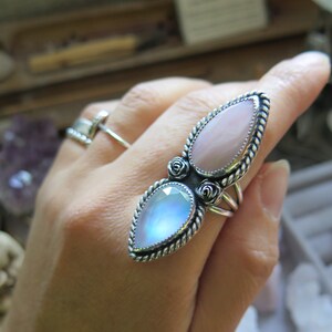May include: A silver ring featuring two teardrop-shaped stones. The top stone is a pale pink, and the bottom stone is an iridescent blue. The ring is adorned with small silver roses.