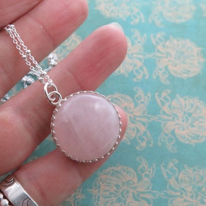May include: A silver necklace with a large, round, pale pink rose quartz pendant. The pendant is set in a decorative silver bezel. The necklace chain is a delicate silver beaded style, suitable for daily wear.