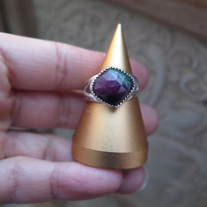May include: A silver ring featuring a large, multi-coloured, faceted gemstone. The gemstone shows shades of purple, green, and blue. The ring is displayed on a gold-coloured, cone-shaped stand.