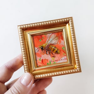 May include: A small framed painting of a bee on a red flower. The bee is yellow and black with brown wings. The painting is in a gold frame with a decorative border.