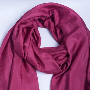 May include: A draped, solid-colored scarf in a deep magenta hue. The fabric appears to be silk or a similar lightweight material, with a subtle sheen. The scarf is arranged in soft folds, creating a flowing, elegant look. The scarf is suitable for all seasons.