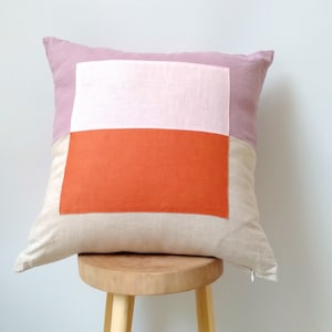 Könnte beinhalten: Ein Dekokissen mit geometrischem Muster in den Farben Rosa, Orange und Beige. Das Kissen ist aus Leinen gefertigt und hat eine schlichte, moderne Optik.
