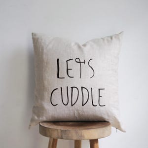 Puede incluir: Una almohada de lino beige con texto negro que dice "Let's Cuddle". La almohada está sentada en un taburete de madera.