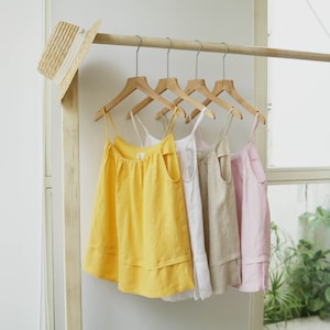 Könnte beinhalten: Vier ärmellose Oberteile hängen an Holzbügeln. Die Oberteile sind gelb, weiß, beige und rosa. Die Oberteile sind aus einem leichten Stoff gefertigt und haben einen Rüschen-Saum.