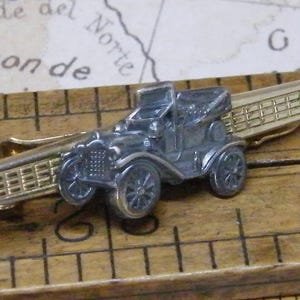 May include: A vintage silver-colored car tie clip with a gold-tone clip. The car is a classic model, likely from the early 20th century. The clip is resting on a wooden ruler with inch markings, and a map is in the background.