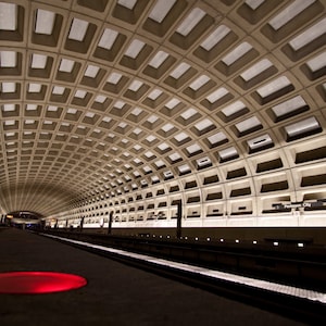 May include: A view of a subway station with a tiled ceiling and a platform with a red circle on the floor. The station is lit with fluorescent lights and has a sign that reads "Pentagon City".