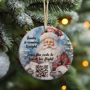 May include: Circular Christmas tree decoration depicting Santa Claus in a red suit and white beard, holding a paper with a QR code. The ornament has the text "Santa is coming tonight" and "Scan the code to track his flight". It hangs from a gold string.
