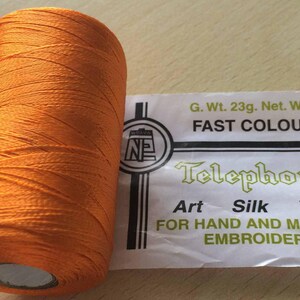 Op de afbeelding: Een spoel oranje zijden draad voor hand- en machineborduurwerk. Het etiket vermeldt "Telephone Art Silk Yarn" en "Fast Colour". De spoel weegt 23 gram en heeft een nettogewicht van 19 gram.
