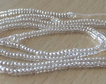 May include: A strand of silver seed beads, approximately 10 feet long, is coiled on a wooden surface. The beads are small and uniform in size, creating a smooth, continuous line.