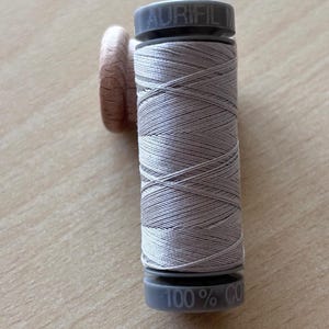 Puede incluir: Un carrete de hilo gris claro con la marca "AURIFIL" impresa en la parte superior. El carrete es de plástico gris y el hilo está enrollado alrededor de un núcleo de madera. La parte inferior del carrete dice "100% CO".