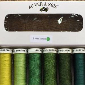 Puede incluir: Una caja de seis carretes de hilo de bordar Au Ver A Soie Surfine en tonos verdes y amarillos. La caja es blanca con el nombre de la marca y la información del producto impresa en ella.