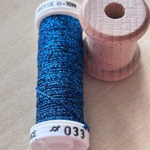 Può includere: Una bobina di filo blu metallizzato con macchioline nere, dotata di tappi in plastica bianca con le scritte TRESSE 8-10M e 033, posizionata accanto a una bobina di legno vuota con centro cilindrico ed estremità svasate e venature visibili.