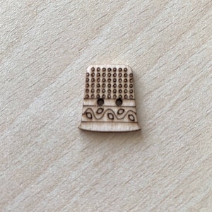 Peut inclure: Un bouton en bois en forme de dé à coudre avec un motif décoratif de feuilles et un motif à pois.