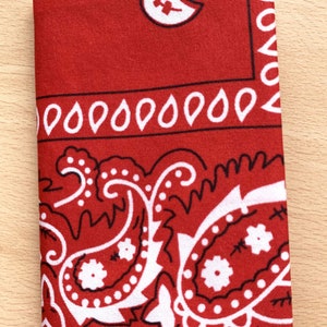 Peut inclure: Un bandana rouge avec un motif paisley blanc. Le bandana a une bordure noire et un petit motif paisley blanc dans le coin supérieur droit.
