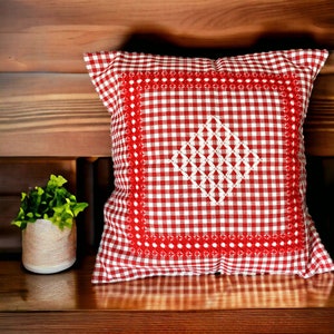 Peut inclure: Un coussin carré décoratif à motif vichy rouge et blanc. Le coussin présente un motif losange brodé blanc au centre et une bordure brodée rouge. Il est posé sur une surface en bois.