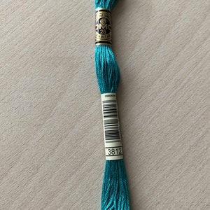 Puede incluir: Madeja de hilo de bordar DMC 25 en color turquesa vibrante. La etiqueta indica el número de color 3812 y una longitud de 8 m (8,7 yd). El hilo está bien enrollado, listo para proyectos de bordado y costura.