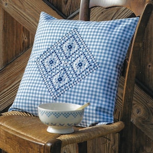 May include: A blue and white gingham cushion with a blue embroidered design sits on a wooden chair with a woven seat. A white bowl with a blue design sits on the chair.