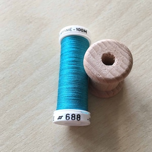 May include: A spool of bright turquoise thread and a wooden bobbin sit on a light wood surface. The thread spool has the number 688 printed on it. The bobbin is made of light-colored wood and has a hole in the center.