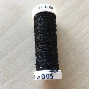 Puede incluir: Una bobina de hilo metálico negro con una etiqueta blanca que dice "TRESSE 8-10M" y "005".