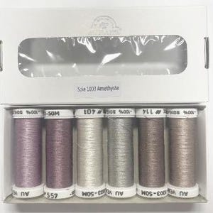 Puede incluir: Un conjunto de seis carretes de hilo de bordar en tonos de morado, blanco y gris. Los carretes están etiquetados "Soie 1003 Amethyste".