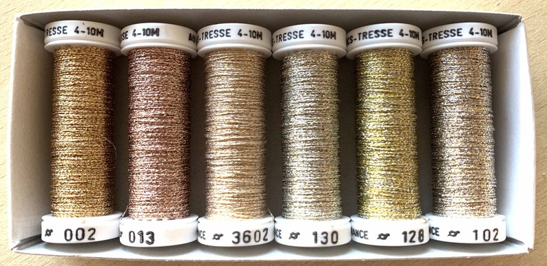 Op de afbeelding: Een doos met zes spoelen metallic garen in goud, zilver en ros&eacute; goud. De spoelen zijn genummerd: 002, 013, 3602, 130, 128 en 102.