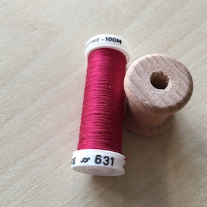 May include: A spool of red thread with a white label that says "# 631" and "FINE - 100M". The spool is sitting on a wooden bobbin.