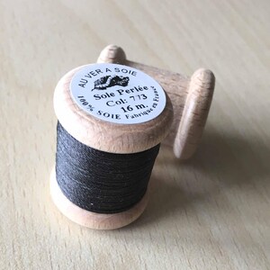 Puede incluir: Una bobina de hilo negro etiquetada "Sole Perlée" con el texto "100% SOIE Fabrique en France" y "Col: 773 16 m."