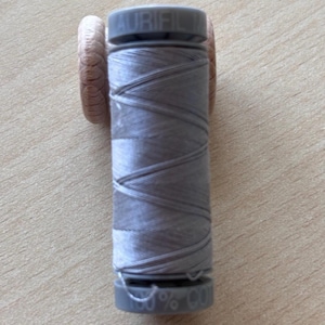 Puede incluir: Un carrete de hilo Aurifil gris claro. El hilo está enrollado alrededor de un carrete de plástico gris, con dos soportes de hilo de madera a cada lado. El texto "AURIFIL" está impreso en la parte superior del carrete. Ideal para coser.