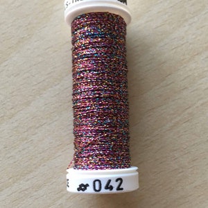 Può includere: Un'bobina di filo da ricamo metallizzato in tonalità rosa, oro e turchese. La bobina è etichettata "TRESSE 4-10M #042".