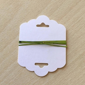 Peut inclure: Carte blanche avec des bords festonnés et deux trous en haut et en bas. Une ficelle métallique verte est enroulée autour de la carte.