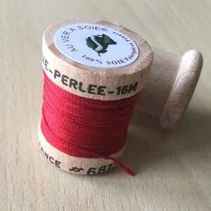 Può includere: Un'bobina di filo rosso con un'rocchetto di legno. L'etichetta sull'rocchetto dice "Au Vera Soie 100% Soie Fabrique en France - Perlee - 16m # 681".
