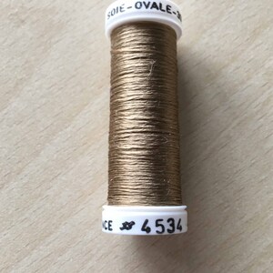 Può includere: Un'bobina di filo dorato con un'etichetta bianca che recita "SOIE - OVALE -" e "# 4534".