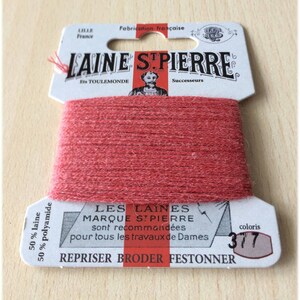 Könnte beinhalten: Eine Spule roten Garns mit der Aufschrift "LAINE STPIERRE" und dem Text "LES LAINES MARQUE STPIERRE sont recommandées pour tous les travaux de Dames" und "REPRISER BRODER FESTONNER" auf dem Etikett. Das Etikett gibt auch an, dass das Garn aus 50% Wolle und 50% Polyamid besteht und die Farbe 317 ist.