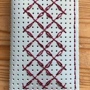 Puede incluir: Una cubierta de cuaderno blanca con una cuadrícula de pequeños agujeros y un patrón cosido en color rosa oscuro. La costura forma un patrón de diamantes en la cubierta, con un borde a juego.