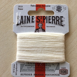 May include: A spool of white embroidery floss with a label that reads "LAINE ST PIERRE" and "Ets TOULEMONDE Successeurs". The label also states "Fabrication française" and "50% laine 50% polyamide". The label has a red border and the text "LES LAINES MARQUE ST PIERRE sont recommandées pour tous les travaux de Dames REPRISER BRODER FESTONNER" is printed in black. The color number "106" is printed in a brown oval.