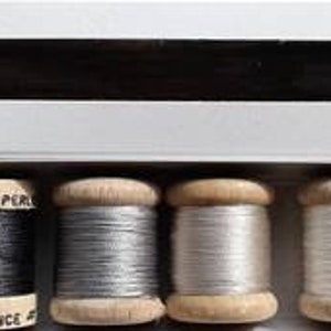 Puede incluir: Un juego de seis carretes de hilo en negro, gris y blanco. Los carretes están etiquetados con "Au Ver A Soie" y "Soie/Silk-France".