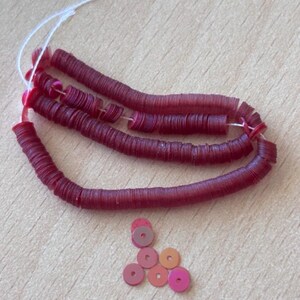 May include: Three strands of dark red, flat, circular clay beads threaded on white string, accompanied by six loose, individual beads in varying shades of red, pink, and orange, each featuring a small, centred hole for stringing.