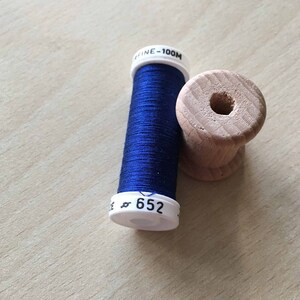 Puede incluir: Una bobina de hilo azul oscuro con una etiqueta blanca que dice "FINE - 100M" y "652". La bobina está sentada sobre una bobina de madera.