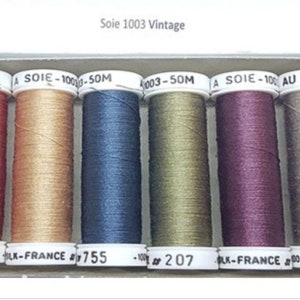 Peut inclure: Six bobines de fil de différentes couleurs, dont le rouge, l'or, le bleu, le vert, le violet et le gris. Chaque bobine est étiquetée "Soie 1003 Vintage" et d'un numéro. Les bobines sont disposées en rangée.