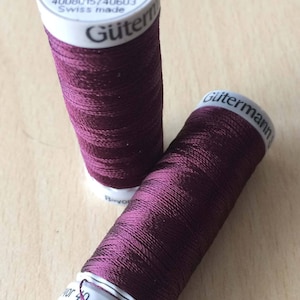 Könnte beinhalten: Zwei Spulen maroonfarbenes Gutermann-Garn. Die Spulen sind mit dem Markennamen und der Farbnummer 1189 gekennzeichnet. Die Spulen sind 200 Meter lang.