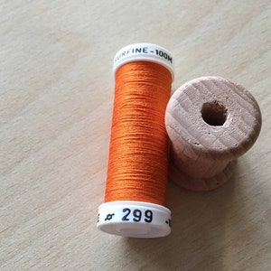 May include: A spool of orange thread labeled "SURFINE - 100M" and "# 299" with a wooden thread bobbin.